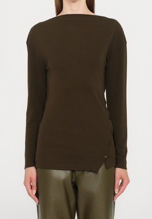 Femme portant un haut à manches longues marron foncé avec des détails de boutons sur le bas côté droit, associé à un pantalon vert olive.