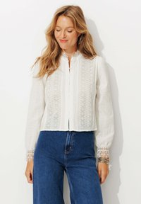 Blouse blanche avec un tissu texturé et des détails à motifs verticaux, col montant, devant à boutons et accents en dentelle aux poignets.