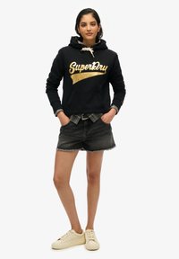 Sudadera negra con capucha y logo dorado, camisa de cuadros debajo, combinada con pantalones cortos de mezclilla negros y zapatillas beige. Atuendo simple y casual.
