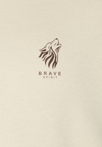 Beige pamut ing, amelyen sötétbarna farkas grafika található, alatta a "BRAVE SPIRIT" felirattal, puha tapintással és laza szabással.