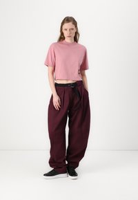Rosa kort topp med korte ermer og en svart logo, kombinert med løstsittende burgunderbukser med svart belte og lommer.