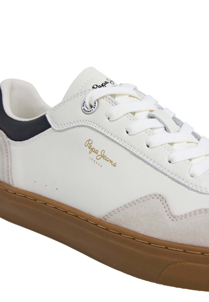 Weißer Sneaker aus Wildleder und synthetischen Materialien mit einer braunen Gummisohle, weißen Schnürsenkeln, goldenem Logo und einem schwarzen Akzent an der Ferse.