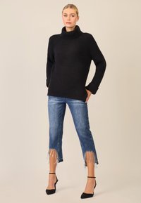 Maglione nero a coste con colletto alto, abbinato a jeans in denim blu con orli frangiati decorati con perline. Scarpe nere con tacco completano il look.
