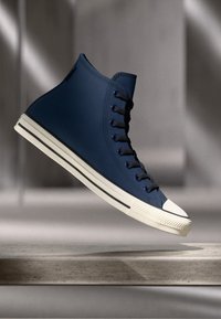 Hoge marineblauwe canvas sneaker met zwarte veters en een witte rubberen neus. Heeft een gemêleerd oppervlak en contrasterende stikseldetails.