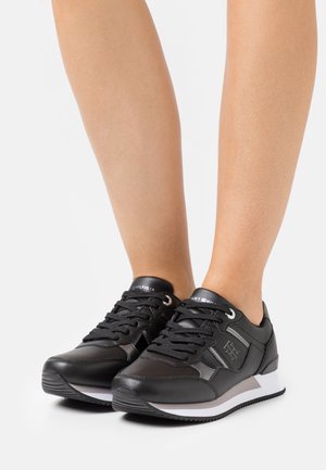 Schwarze Sneaker mit einer glatten synthetischen Oberseite, Schnürdesign, weißer Zwischensohle und Logo-Akzenten auf der Zunge und der Seite.