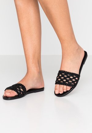 Nohy obuté v černých tkaných sandálech typu slip-on s otevřenou špičkou a plochou podrážkou na světle podlaze s jednoduchým světlým pozadím.