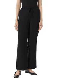 Schwarze, weite Hosen aus einem leichten Stoff, mit einem elastischen Bund und einer Kordel sowie zwei Seitentaschen. Flache Schuhe sind sichtbar.