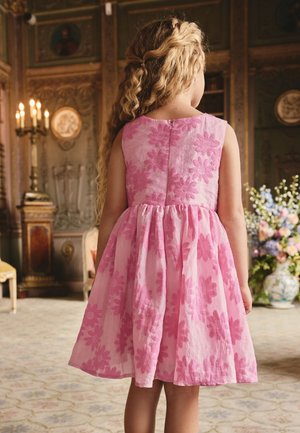 Pige med langt krøllet hår iført en ærmeløs lyserød kjole med blomsterprint står i et udsmykket rum med vintage dekoration.