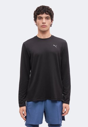 Ung mand iført en sort langærmet Puma trøje og blå atletikshorts, stående mod en ensartet lys baggrund.