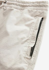 Shorts beige chiaro realizzati in tessuto a trama. Presentano una vita elasticizzata, una tasca laterale con zip e hardware nero, e dettagli cuciti.