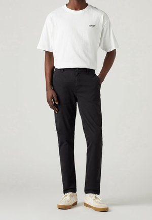 Homme portant un t-shirt blanc Levi's, un pantalon noir et des baskets blanches, debout avec une main dans la poche devant un fond gris clair.