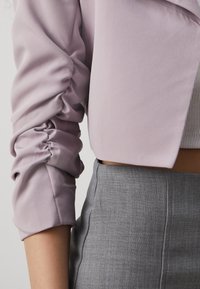 Chaqueta de color púrpura claro, con mangas abullonadas en tejido suave, ceñida en la muñeca. Combinada con una falda gris ajustada, que muestra un diseño limpio y estructurado.