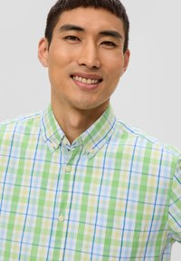 Camicia a maniche corte con colletto, caratterizzata da un motivo a quadri blu e verde con accenti bianchi, chiusura frontale con bottoni e una texture di tessuto leggera.
