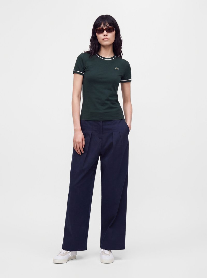 Femme portant un t-shirt ajusté vert foncé, un pantalon large bleu marine, des baskets blanches et des lunettes de soleil, debout les mains dans les poches sur un fond uni.