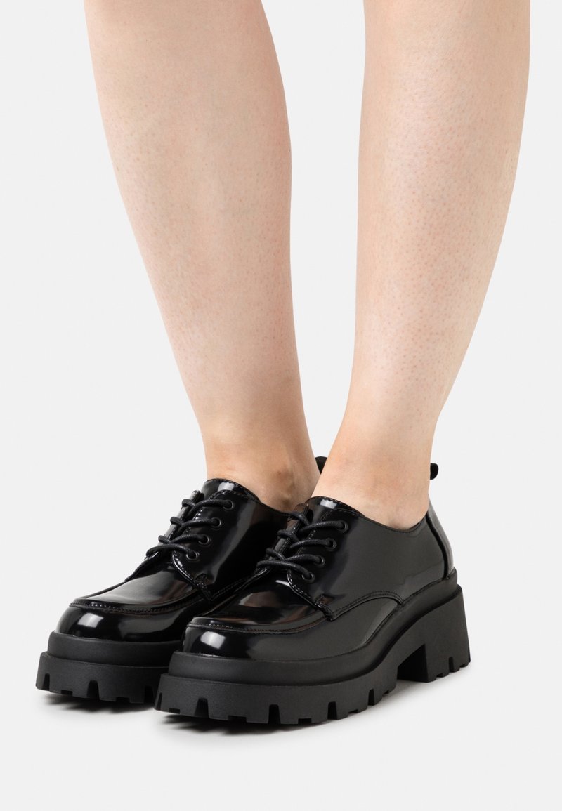 Chaussures à plateforme épaisses noires brillantes à lacets portées sur des chevilles nues, mettant en valeur des semelles épaisses avec une profonde bande de roulement et une finition polie.