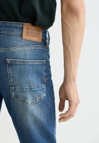Vaqueros de denim azul medio con una parche de cuero etiquetado "SCOTCH & SODA AMSTERDAM". Presentan doble costura y una pequeña puntada de acento amarilla.