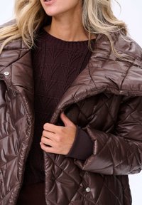 Brązowa pikowana kurtka puchowa z wysokim kołnierzem i zapięciem na napy, noszona na ciemnofioletowym swetrze o teksturowanym splocie. Gładkie wykończenie materiału.