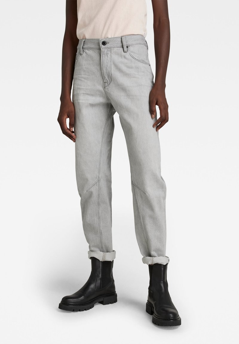 Personne portant un jean gris clair retroussé aux chevilles et des bottines en cuir noir, debout devant un fond uni.