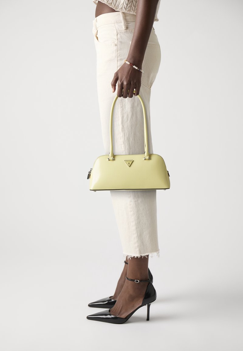 Personne portant un jean crème court et des talons hauts noirs, tenant un sac à main jaune clair par ses poignées sur le côté.