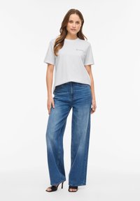 Γυναίκα που στέκεται φορώντας λευκό κοντομάνικο μπλουζάκι με τη λέξη "amore" και ψηλόμεσο παντελόνι με φαρδιά μπατζάκια σε μπλε χρώμα, συνοδευόμενο από μαύρες σανδάλια με τακούνι.