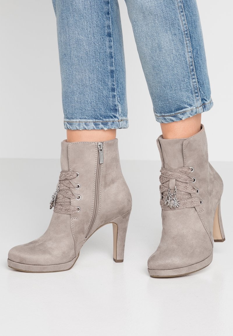 Bottes à talons en daim taupe jusqu'à la cheville avec fermetures éclair latérales, lacets à l'avant, breloques décoratives, portées avec un jean denim bleu court.