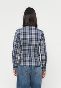 Person med skulderlangt mørkt hår iført en blå og hvit rutete skjorte og blå jeans, som står vendt mot en enkel hvit vegg.