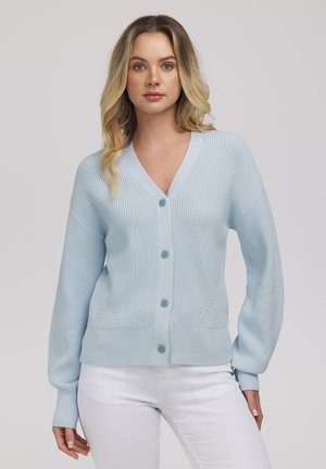 Kobieta w jasnoniebieskim, zapinanym na guziki kardiganie o prążkowanej fakturze i białych spodniach, stojąca na tle gładkiego, jasnoszarego tła.