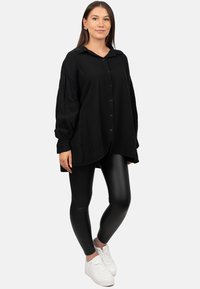 Camicia nera oversize con bottoni, in tessuto texturizzato, maniche lunghe e orlo curvo, abbinata a leggings neri lucidi e sneakers bianche.