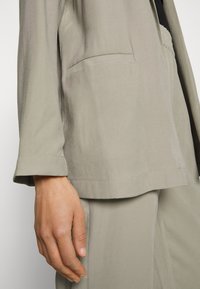 Casaco de manga longa cinza claro, com um bolso. O tecido parece liso e ligeiramente brilhante, apresentando um corte descontraído e detalhes de costura mínimos.