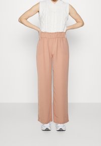 Osoba v bezramenném bílém pleteném topu, volných světle hnědých kalhotách s vysokým pasem a bílých teniskách, stojící s rukama na bocích.