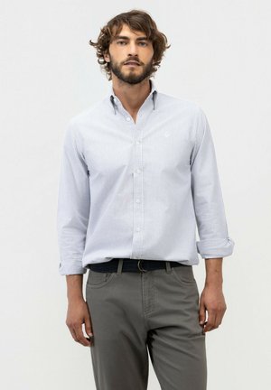 Hombre con cabello rizado y barba, vistiendo una camisa de rayas azul claro abotonada y pantalones grises, de pie frente a un fondo liso.