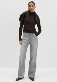 Vrouw in een donkerbruine coltrui met bijpassende bontsjaal, lichtgrijze wijde jeans en puntige bruine schoenen, staand met één hand in de zak.