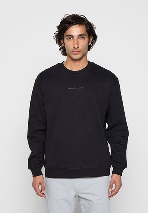 Mężczyzna w czarnej, długorękawowej bluzie z napisem "Marc O’Polo DENIM", w połączeniu z jasnoszarymi spodniami, stojący na jednolitym tle.