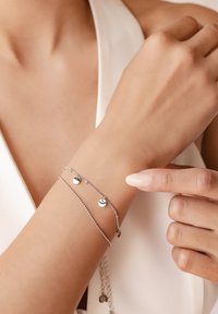 Silver bracelet featuring two small, round charms on a delicate chain, worn on a wrist; hand points toward the bracelet.