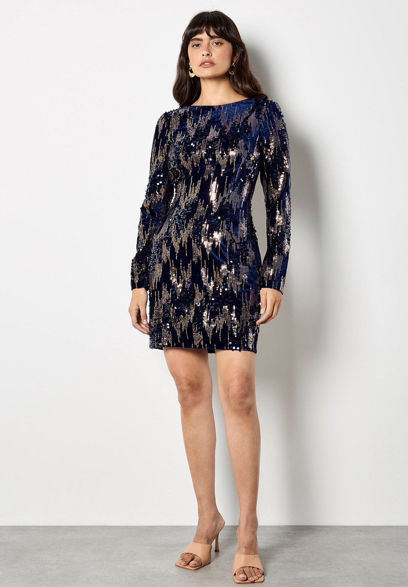 Krótka granatowa sukienka z cekinami z długimi rękawami, z teksturowanym wzorem i dopasowanym krojem. Noszona z cielistymi sandałami na obcasie.