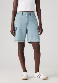 Persona con pantalones cortos cargo de mezclilla azul claro, top corto blanco y zapatillas blancas con rayas rojas y azules, de pie sobre un fondo gris.