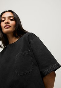 T-shirt en denim noir, coupe décontractée, manches courtes, col rond, avec une poche avant ornée d'un détail de couture visible. Texture lisse.