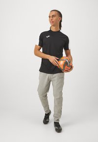 Camisa deportiva negra con mangas blancas, pantaloness cortos grises claros y zapatillas de fútbol negras, sosteniendo un balón de fútbol naranja y negro.
