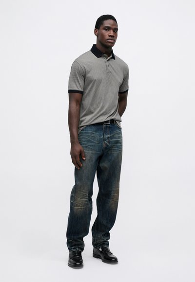 Gråstripet polo skjorte med svart krage, kombinert med falmede blå jeans og svarte dressko, stående mot en ensfarget bakgrunn.