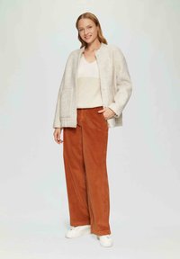 Veste en mélange de laine gris clair avec des fermetures à pression, pull beige à côtes et pantalon large en velours côtelé rouille. Des baskets blanches complètent la tenue.