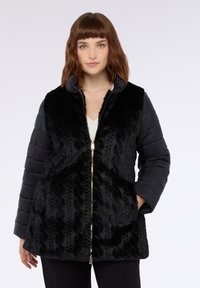 Vrouw met schouderlang bruin haar, gekleed in een zwarte ritsjack met gewatteerde mouwen en een textuur met een vachtachtige voorkant, staand tegen een effen achtergrond.