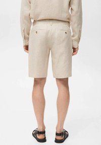 Beige linen shorts with a straight cut, featuring two back pockets with gold accents, paired with black sandals. Flat waistband design.