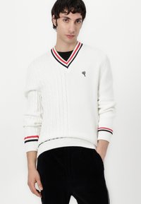 Weißer Strickpullover mit V-Ausschnitt, der auf einer Seite ein Zopfmuster zeigt, schwarze und rote gestreifte Akzente am Kragen und an den Ärmeln sowie ein kleines Herz-Logo.