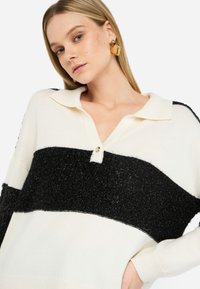Maglione a righe bianche e nere in maglia, con colletto e bottone simile a una perla, caratterizzato da una sezione nera texturizzata e tessuto bianco liscio.