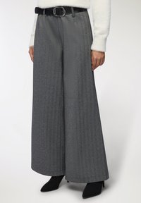 Kobieta w szarych spodni w jodełkę z wysokim stanem i szerokimi nogawkami, z czarnym paskiem, białym puszystym swetrem i czarnych botkach na spiczastym nosku.