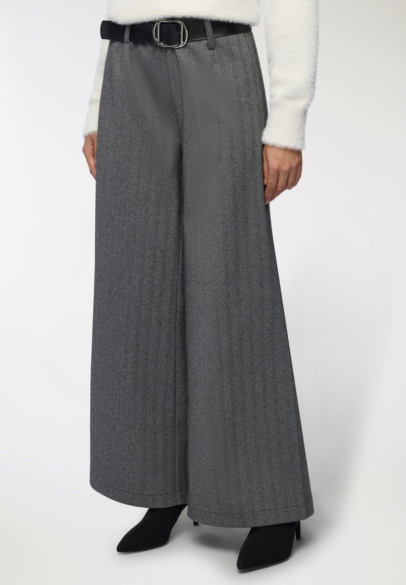 Kobieta w szarych spodni w jodełkę z wysokim stanem i szerokimi nogawkami, z czarnym paskiem, białym puszystym swetrem i czarnych botkach na spiczastym nosku.