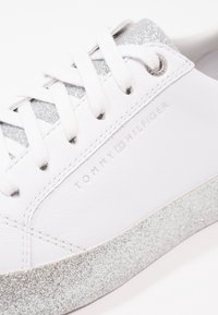 Sneaker bianco in pelle con accento di glitter argento metallico sul tallone, dotato di lacci bianchi e marchio "TOMMY HILFIGER" in rilievo.