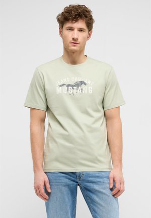 Mann mit lockigem Haar, der ein hellgrünes Mustang-Logo-T-Shirt und blaue Jeans trägt, steht vor einem schlichten hellen Hintergrund.