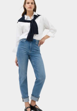 Frau trägt ein weißes Hemd mit Knöpfen und einen dunkelblauen Pullover um die Schultern gebunden, blaue hochgekrempelte Jeans und schwarze Mary-Jane-Schuhe.
