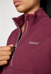 Burgundi színű, cipzáras pulóver magas nyakkal, ezüst hardware-rel és hímzett DKNY logóval. Puha tapintás, kontrasztvarrásos részletekkel.